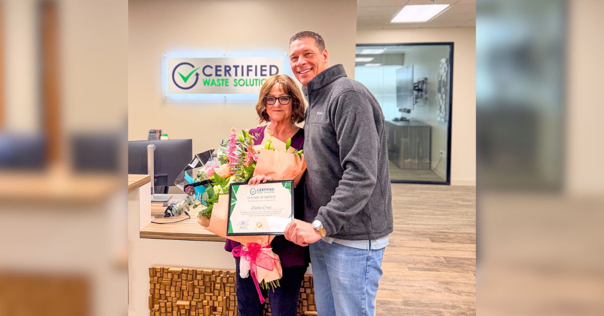 Debbie recognized for 44 years of service at Certified Waste Solutions stands with company leadership holding a certificate and flowers in an office reception area beneath a Certified Waste Solutions sign.